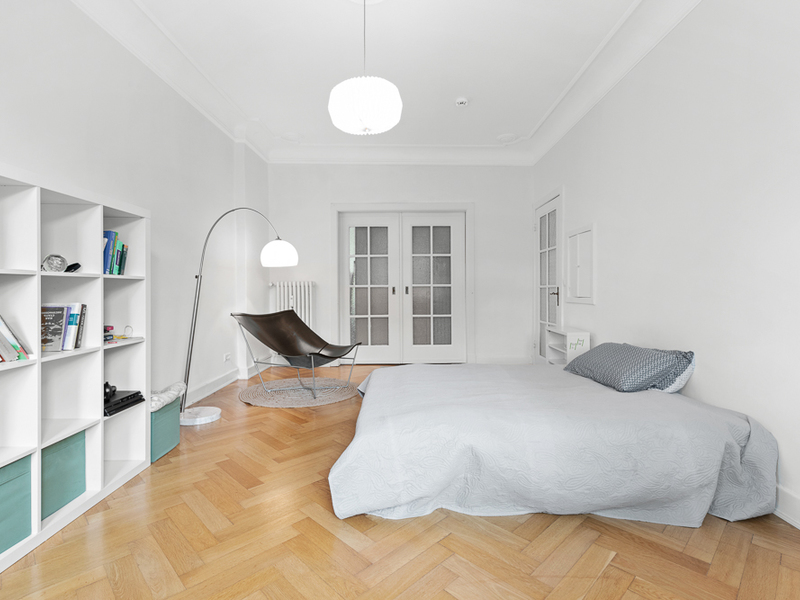 Helles Schlafzimmer mit Doppelbett, Regal, Sessel, Stehlampe und Holzboden.