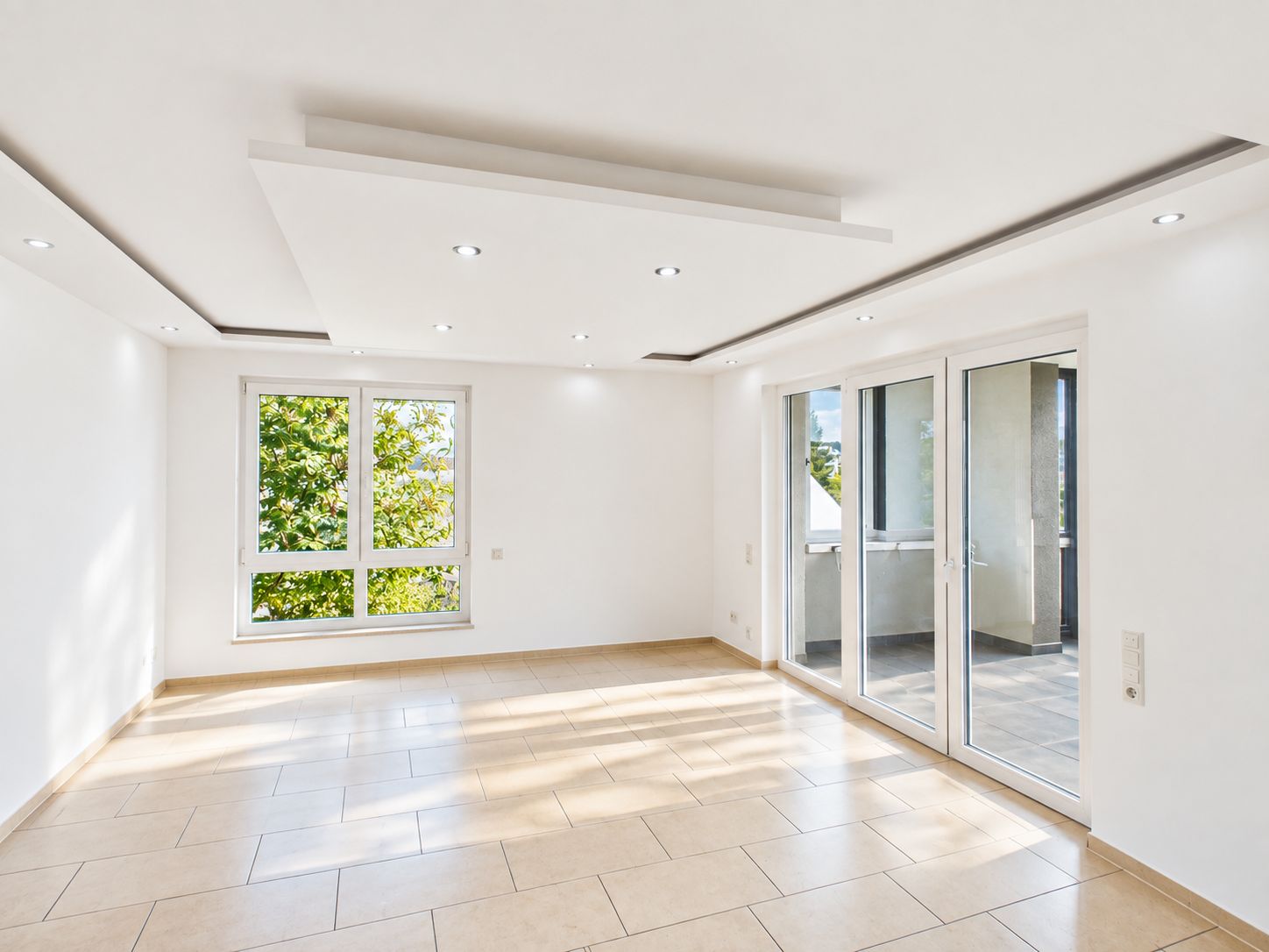 Helles, leeres Wohnzimmer mit großen Fenstern, Fliesenboden und Zugang zu einem Balkon.