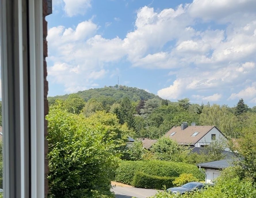 Blick aus dem Fenster auf grüne Hügel, Häuser und einen teils bewölkten Himmel.