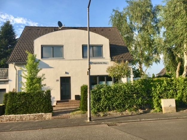 Zweistöckiges weißes Wohnhaus mit dunklem Dach, umgeben von Bäumen und Hecken an einer Straße.