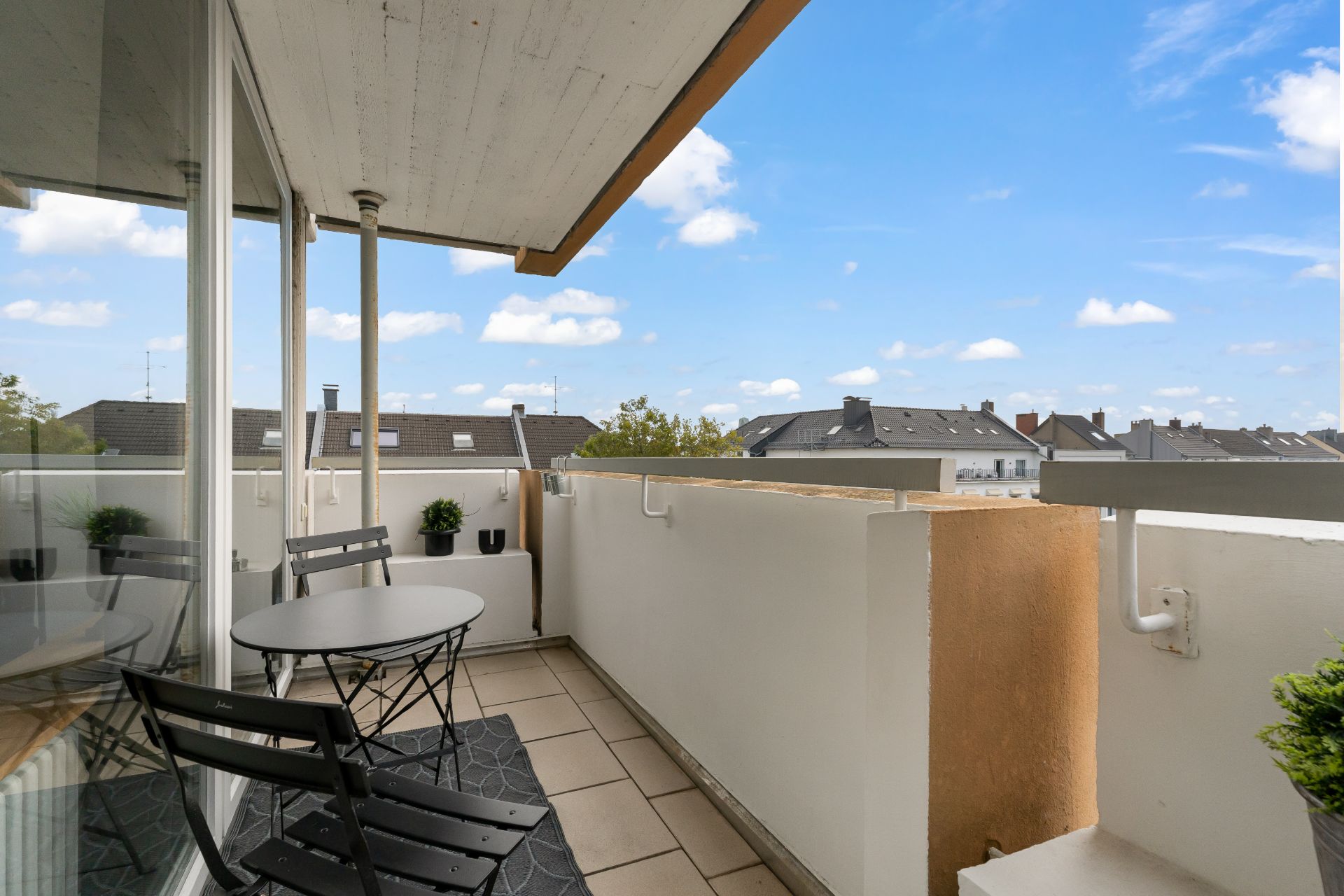 Balkon mit kleinem Tisch, zwei Stühlen und Pflanzen, Blick auf Dächer und blauen Himmel.