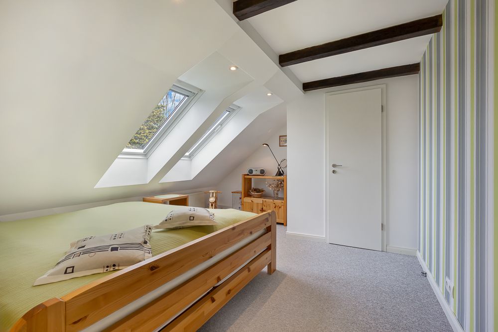 Helles Schlafzimmer mit Dachschrägen, Holzbett, zwei Fenstern und Holzmöbeln.