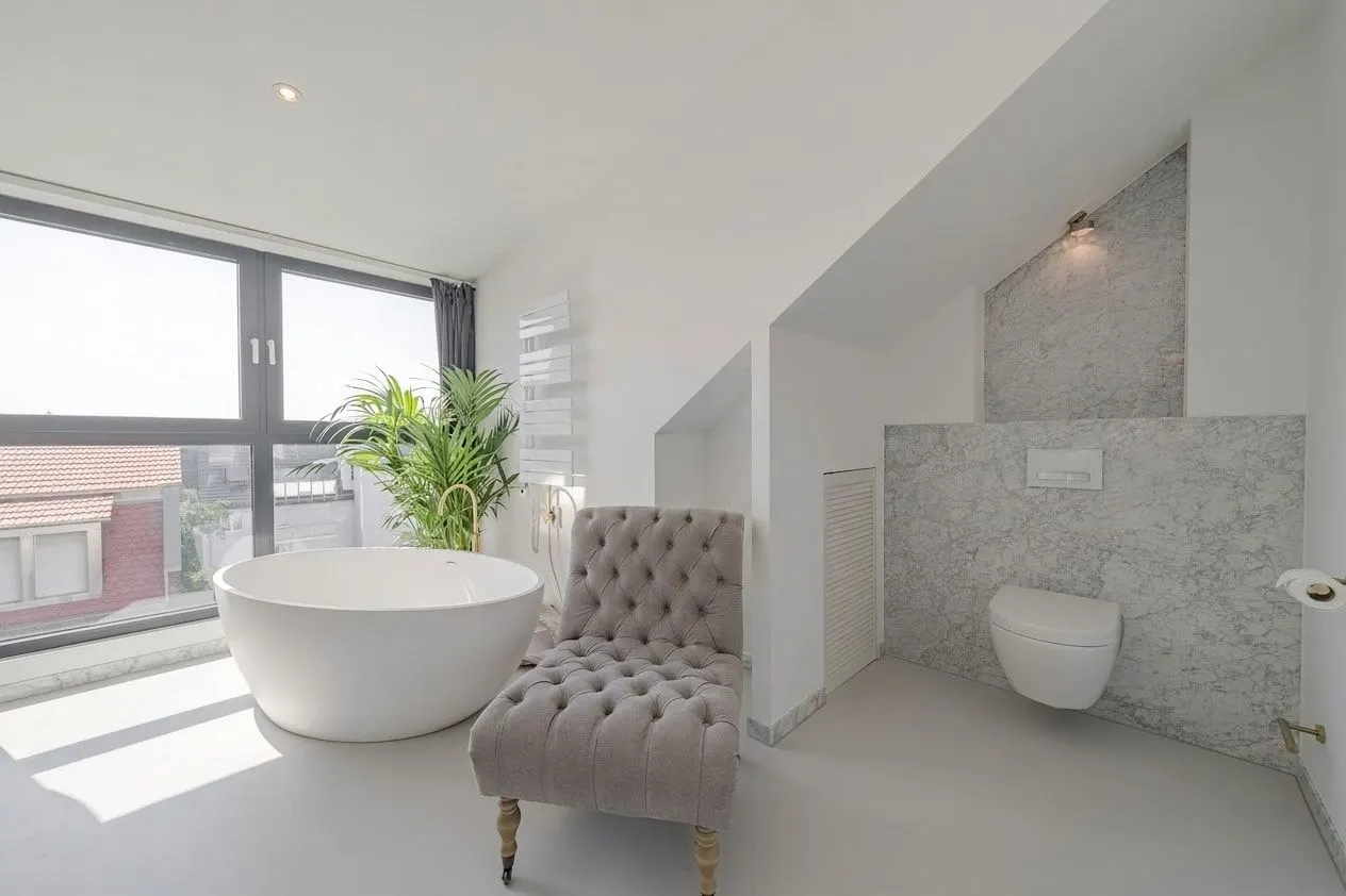 Helles Badezimmer mit freistehender Badewanne, grauem Sessel, großer Fensterfront und WC.