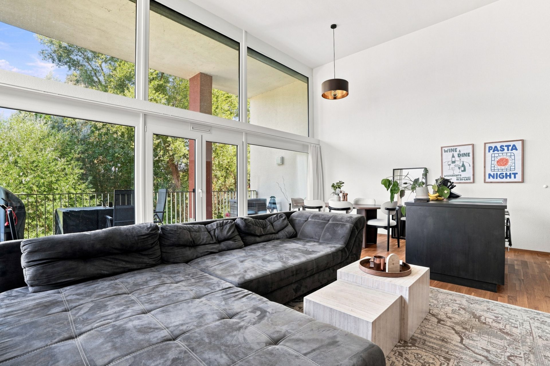 Modernes Wohnzimmer mit großer grauer Eckcouch, Esstisch, Kücheninsel und raumhohen Fenstern mit Blick ins Grüne.