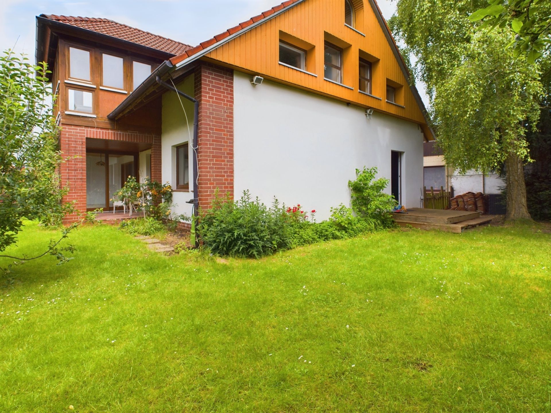 Ein Einfamilienhaus mit rotem Dach, großen Fenstern und Garten mit Rasen und Bäumen.