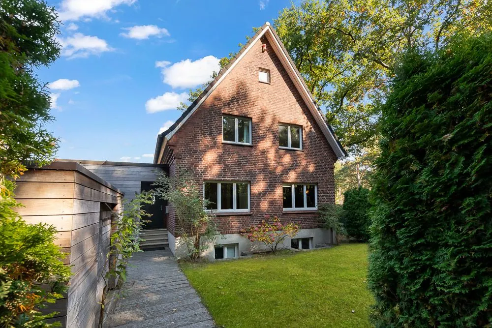 Backsteinhaus mit spitzem Dach, Garten und Gehweg, umgeben von Bäumen und Sträuchern.