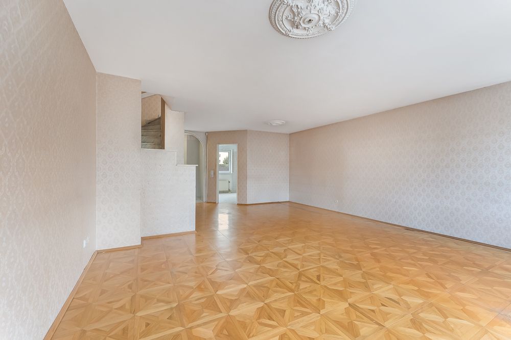 Leeres Wohnzimmer mit gemustertem Parkettboden, hellen Wänden und einer Treppe im Hintergrund.