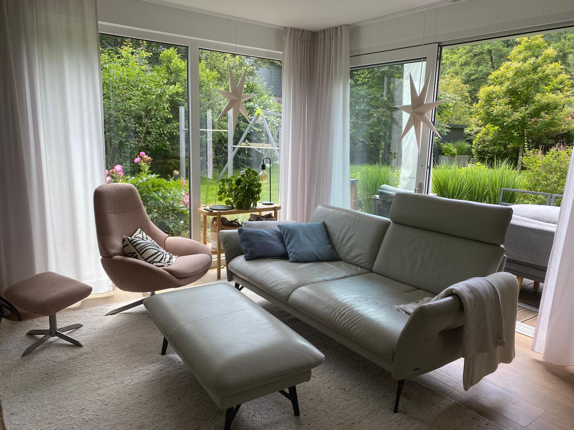 Helles Wohnzimmer mit Sofa, Sessel, Hocker und großen Fenstern mit Blick auf einen Garten.
