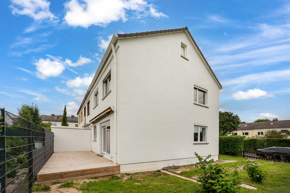 Modernes, weißes Wohnhaus mit Terrasse und Garten, daneben ein Zaun, blauer Himmel im Hintergrund.