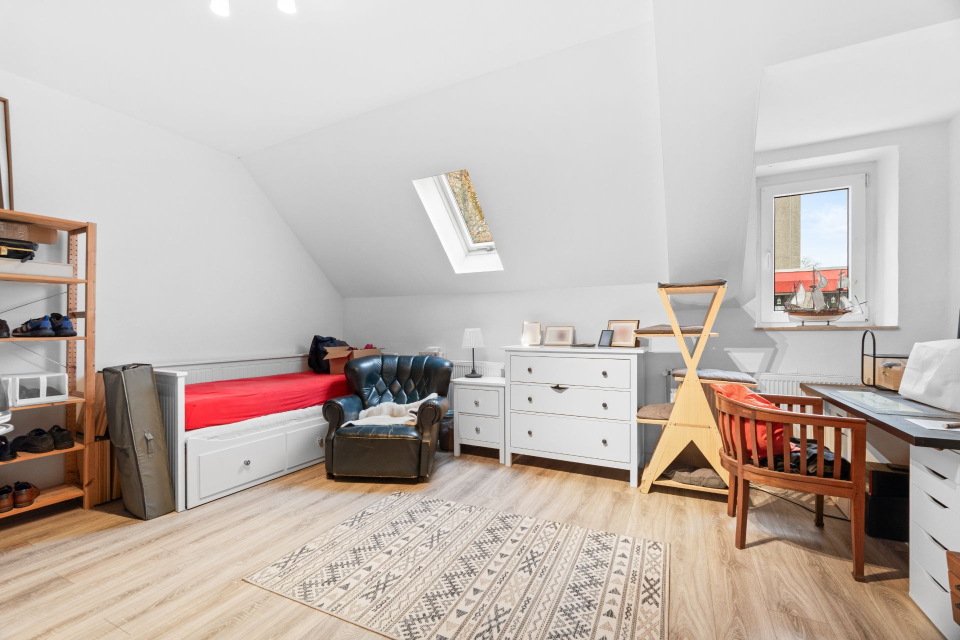 Helles Zimmer mit Einzelbett, Sessel, zwei Kommoden, Schreibtisch und Regal unter Dachschräge.