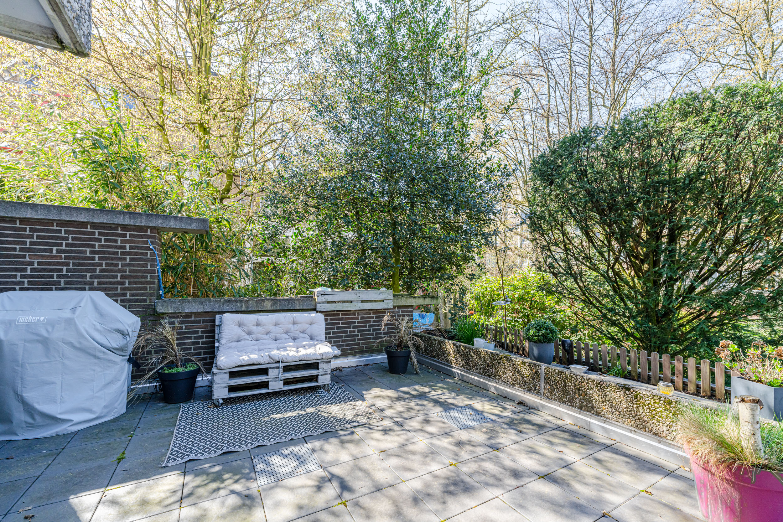 Terrasse mit Bank aus Paletten, Grillabdeckung und vielen Pflanzen, von Bäumen umgeben.