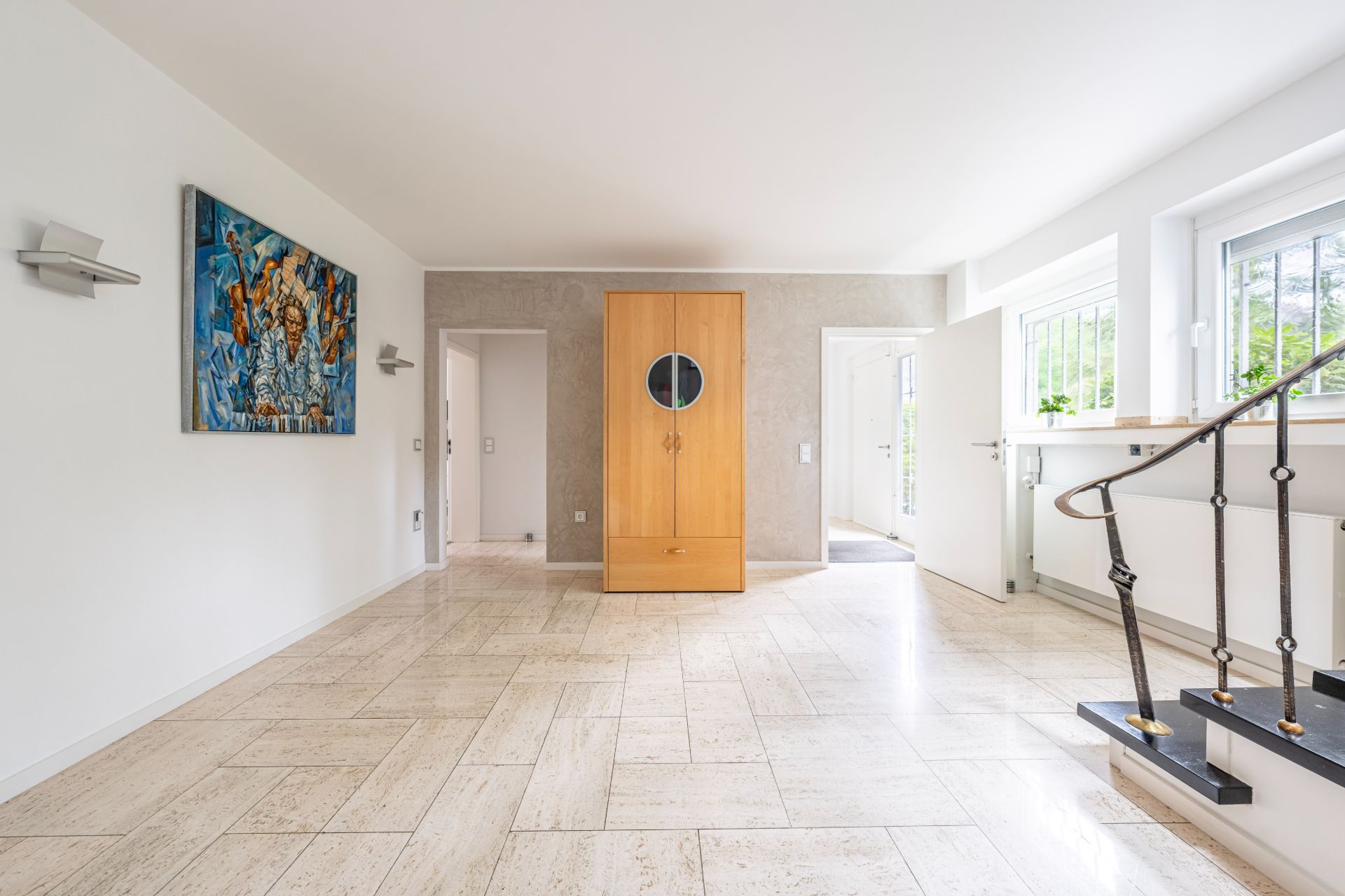 Heller Flur mit Kachelboden, Holzschrank, Treppe, Fenstern und Wandbild.