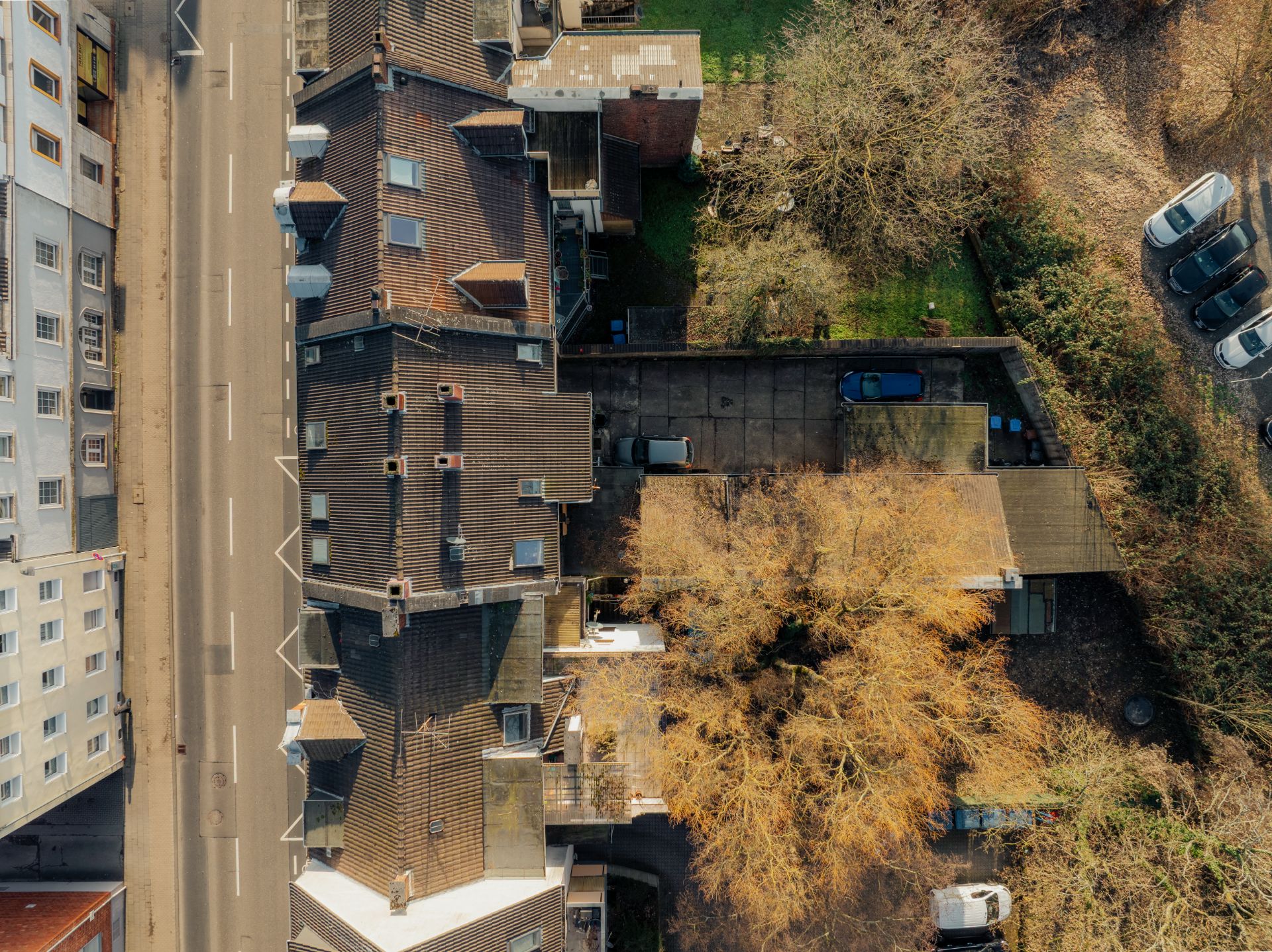 Dachansicht eines Wohnhauses mit Garten, Einfahrt und Bäumen, daneben eine Straße und geparkte Autos.