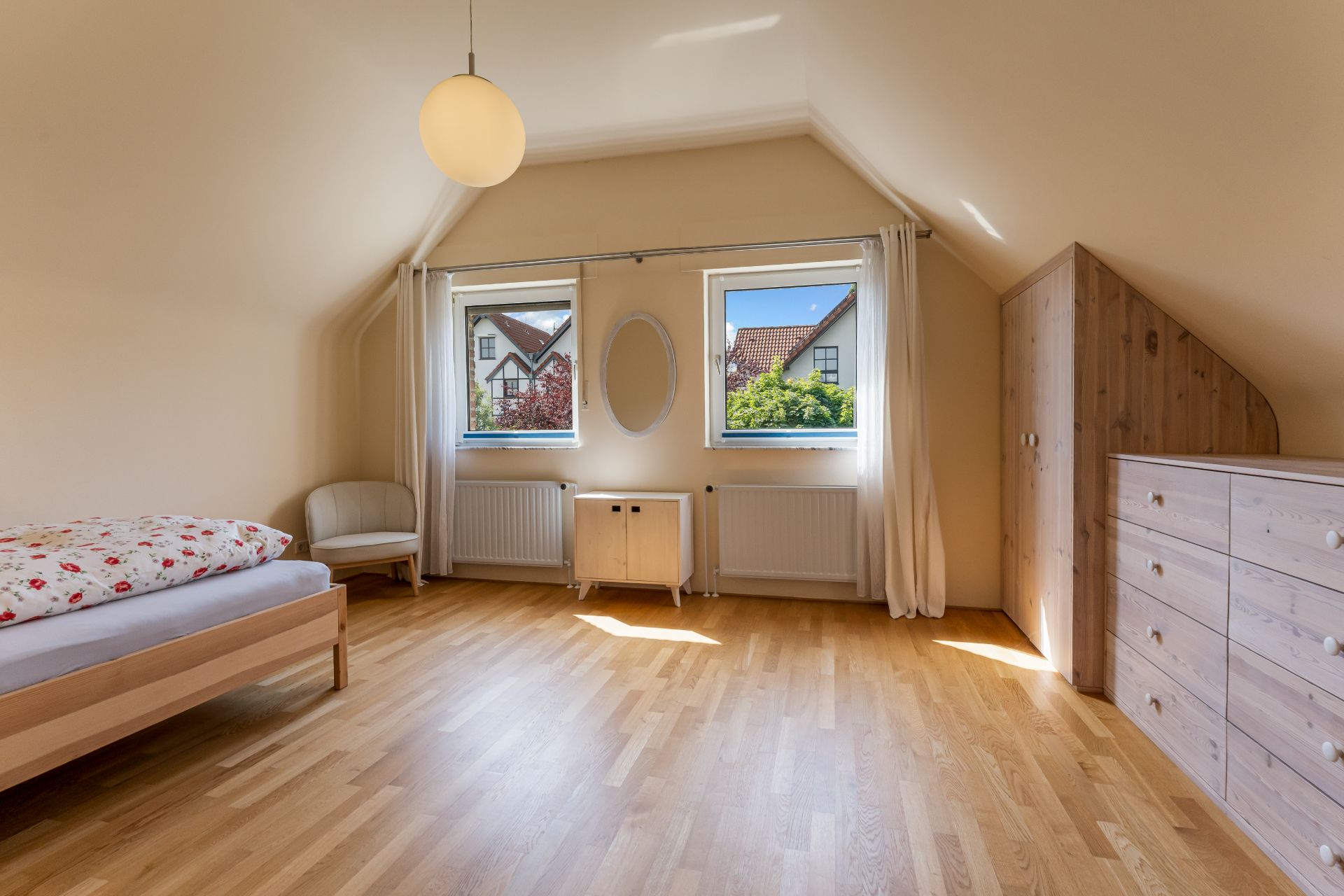 Helles Schlafzimmer mit Bett, Sessel, Kommode und zwei Fenstern mit weißem Vorhang.