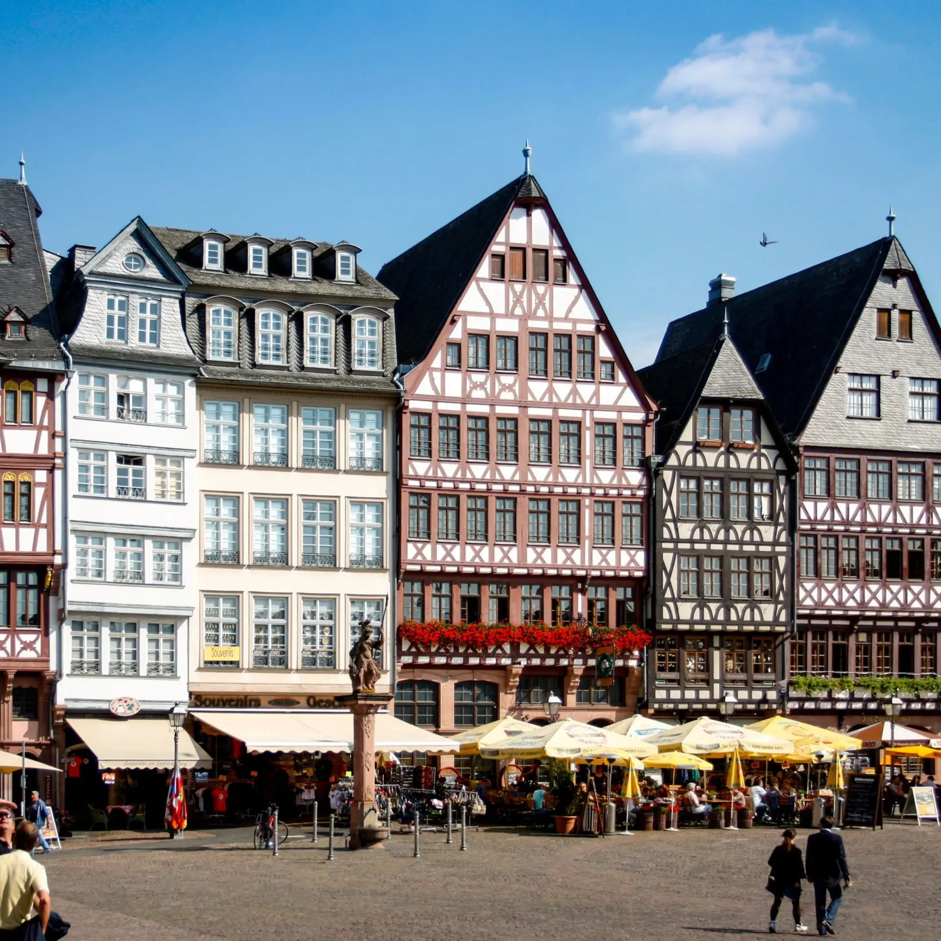 Historische Fachwerkhäuser mit Cafés und Statuen auf einem belebten Platz bei sonnigem Wetter.