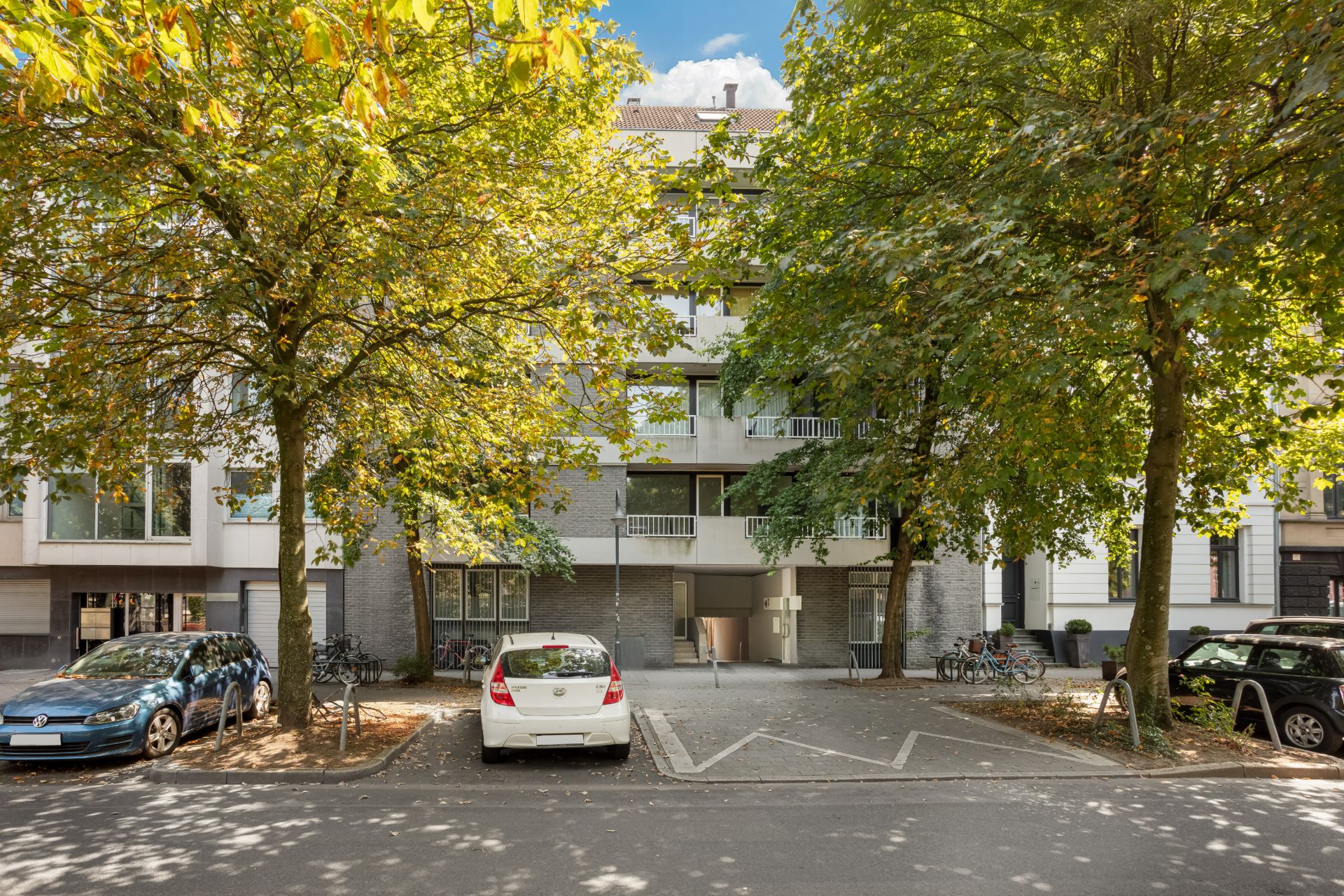 Mehrfamilienhaus mit Balkon und Bäumen, geparkte Autos und Fahrräder vor dem Eingang.