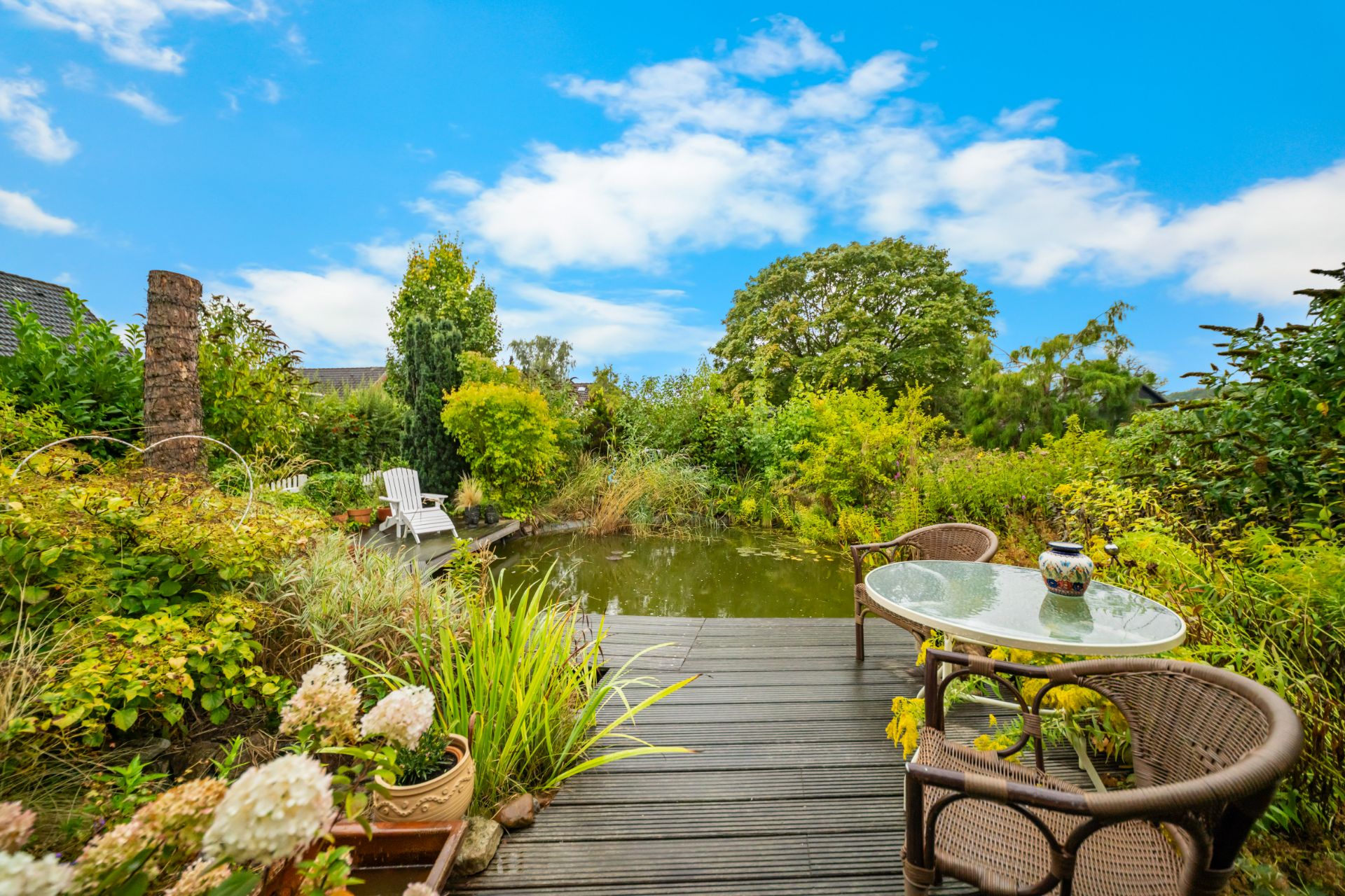 Gartenteich mit Holzterrasse, Tisch, zwei Stühlen und vielen Pflanzen bei blauem Himmel.