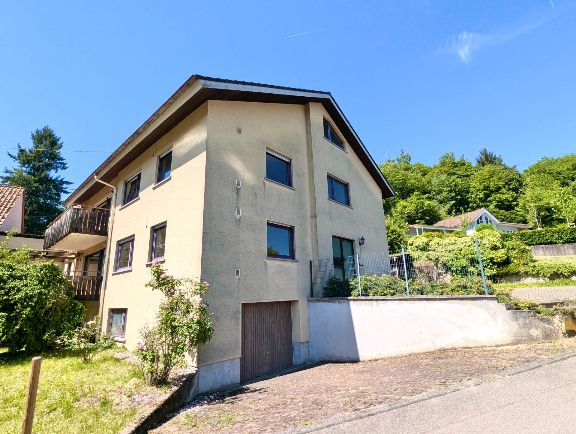 Dreistöckiges Wohnhaus mit Schrägdach, Garage und Balkon, umgeben von Grünpflanzen.