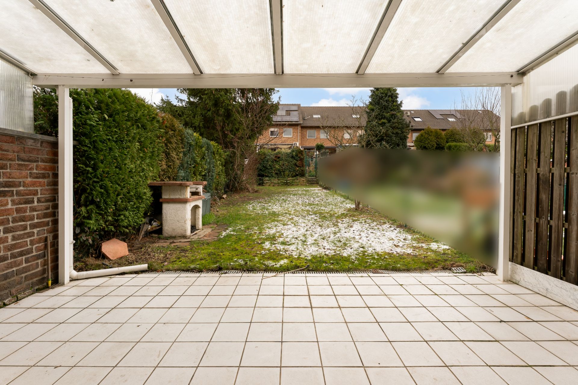 Überdachte Terrasse mit Fliesenboden und Blick auf einen Garten mit Grillplatz.