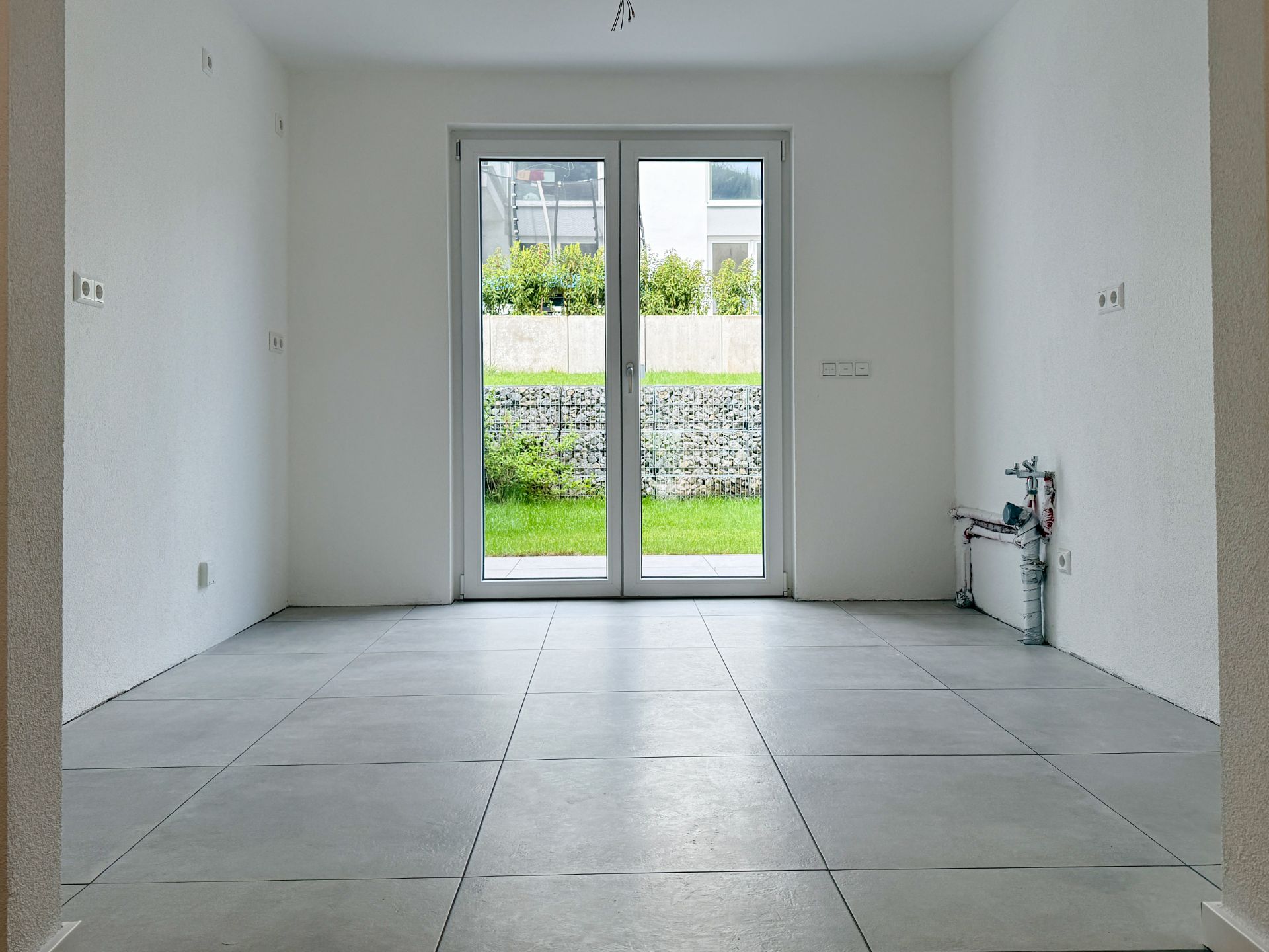 Leerer Raum mit Fliesenboden, großer Glastür und Blick auf einen Garten.