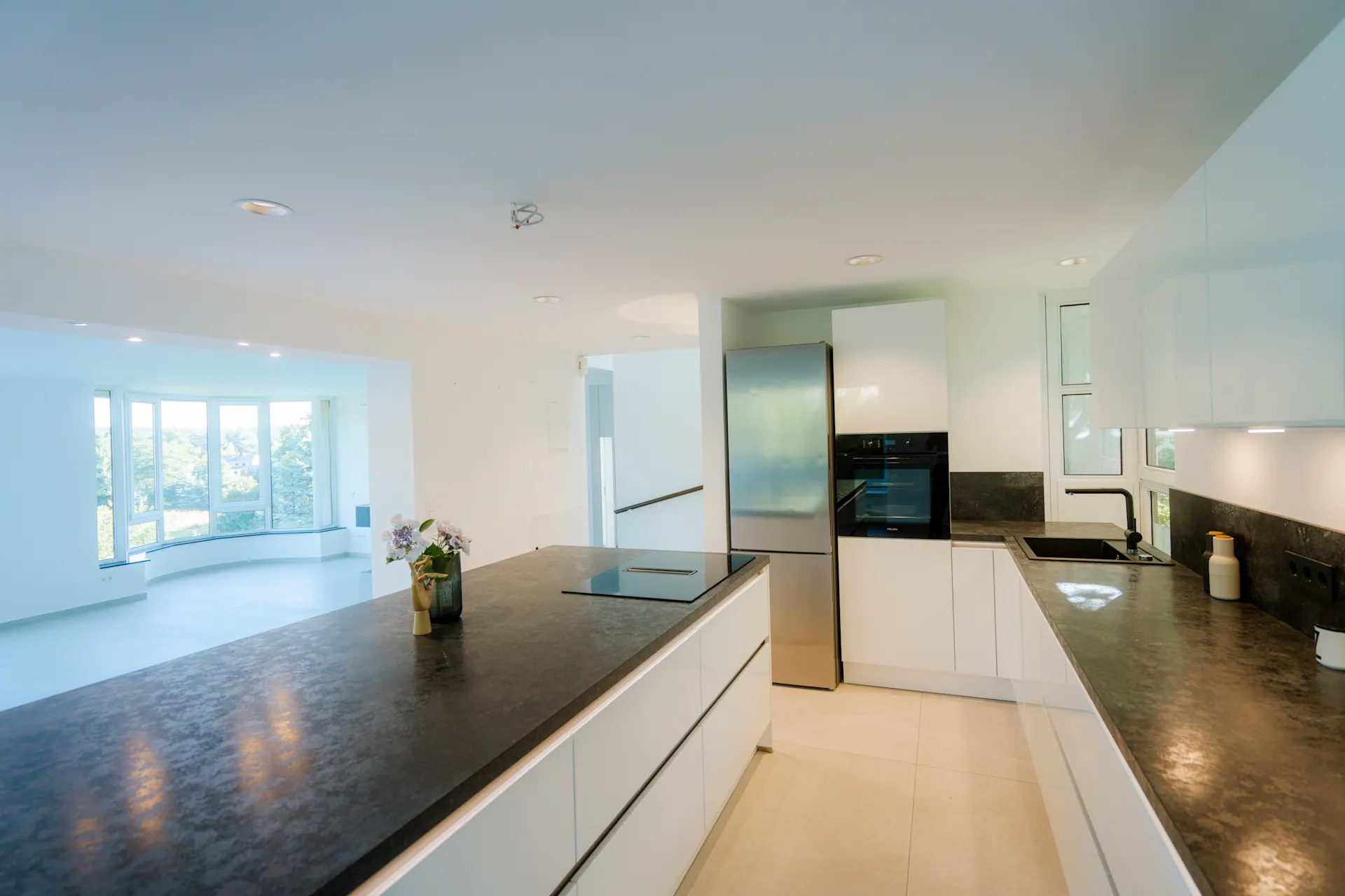 Moderne Küche mit weißen Schränken, Kochinsel und Kühlschrank, angrenzendes helles Wohnzimmer.