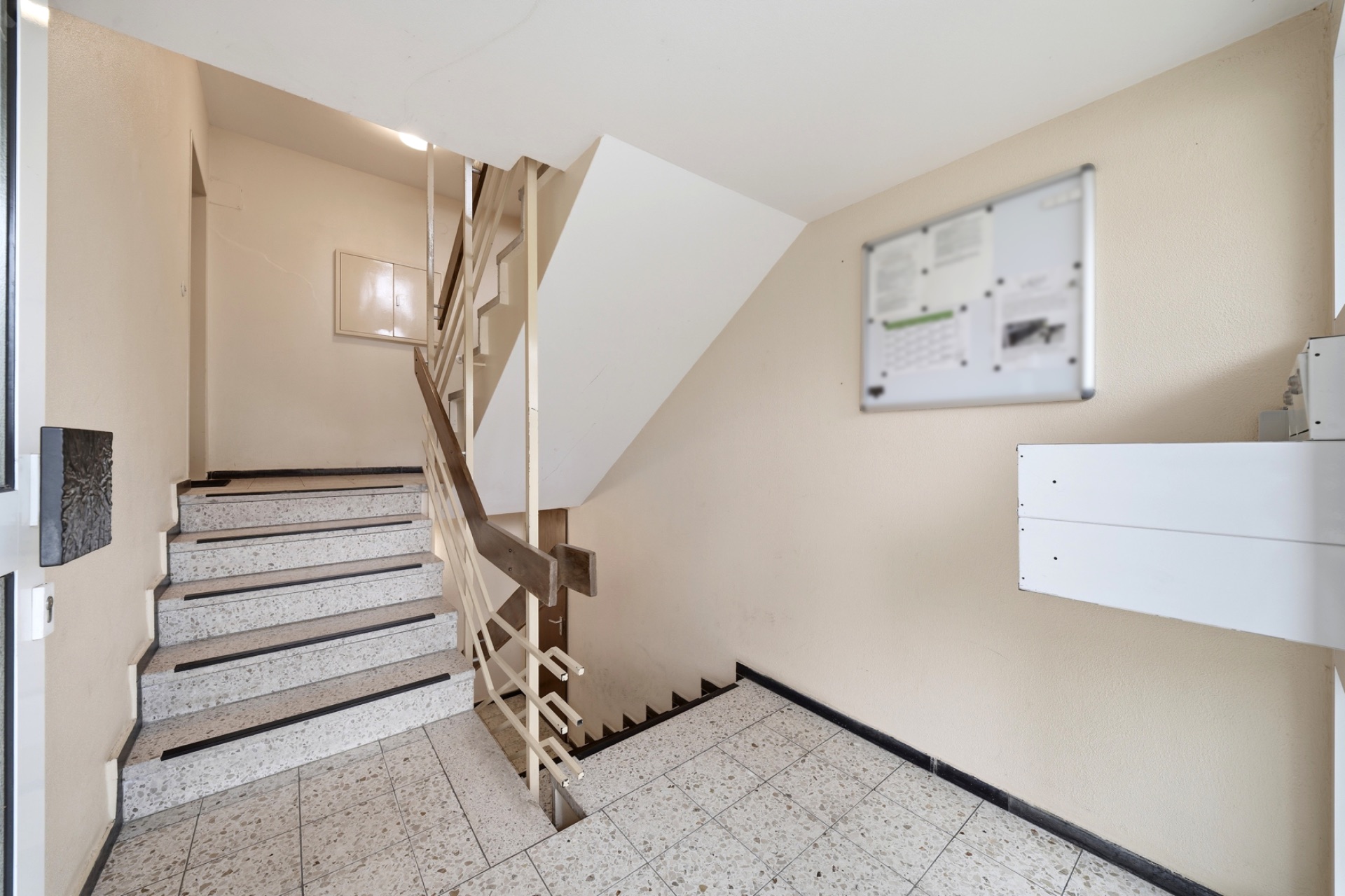 Treppenhaus mit Fliesenboden, Geländer, Infotafel an der Wand und Briefkasten.