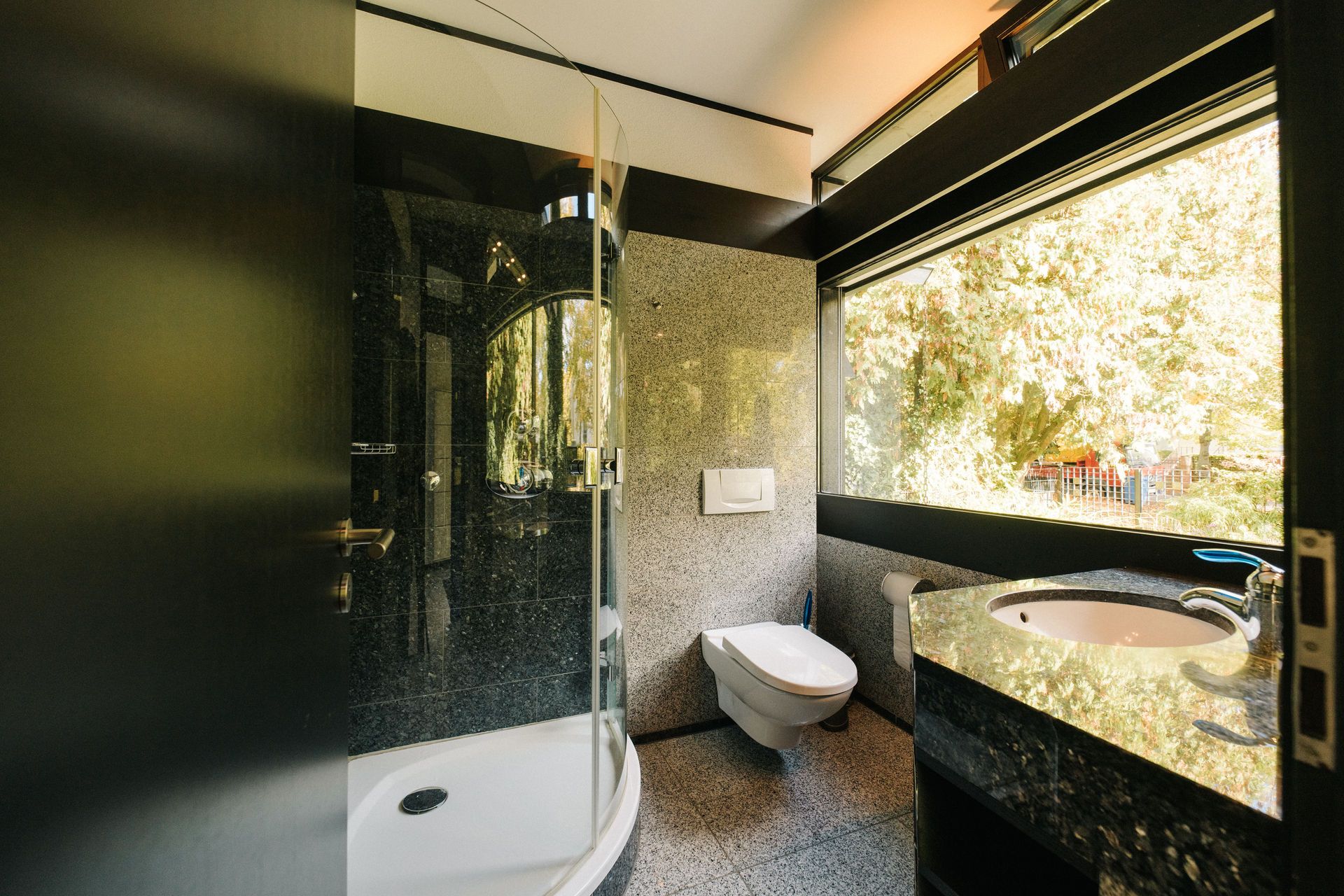 Modernes Badezimmer mit Dusche, Wandtoilette, Waschbecken und großem Fenster mit Blick nach draußen.