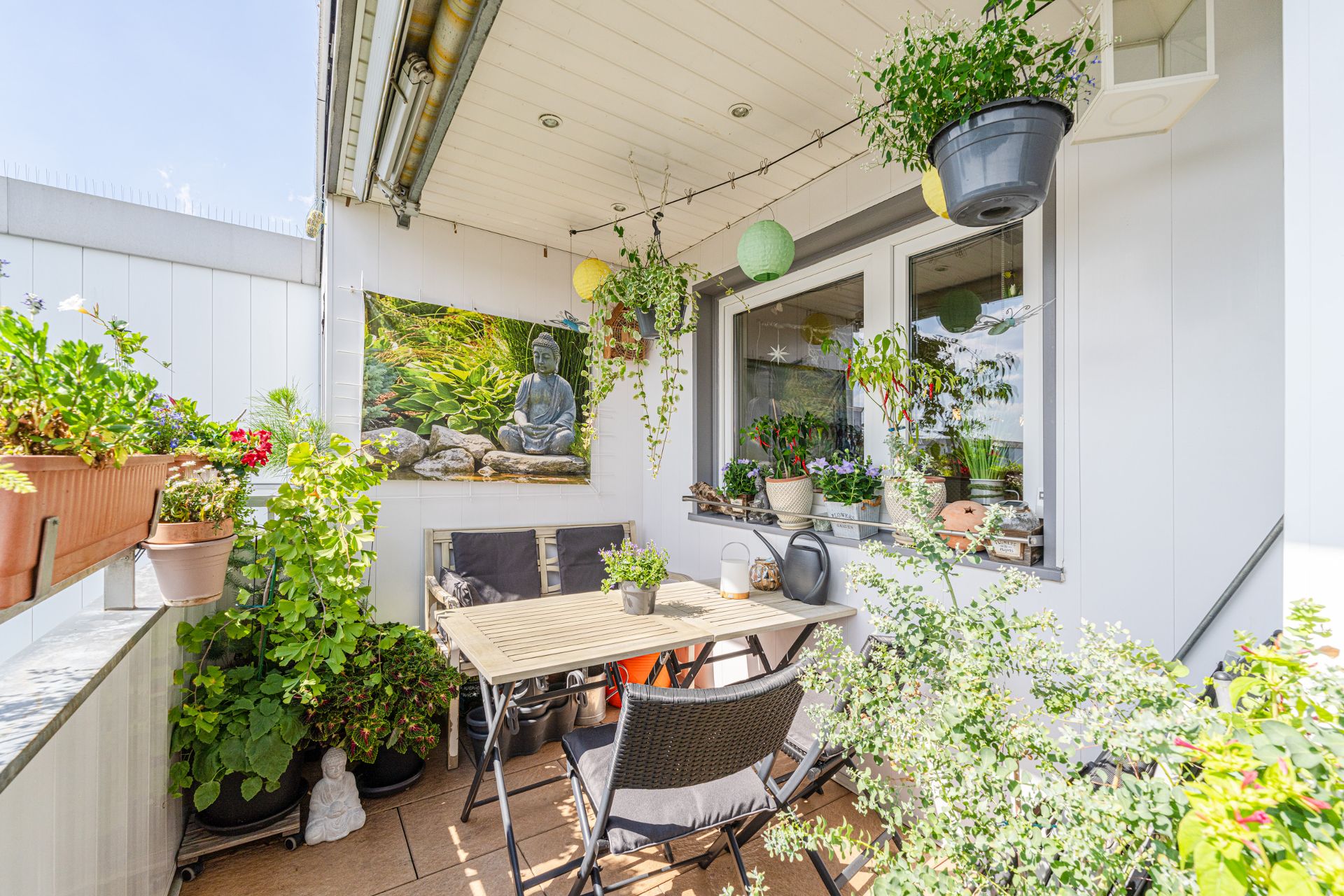 Balkon mit Holztisch, Stühlen, vielen Pflanzen und Wandbild mit Buddha-Figur.