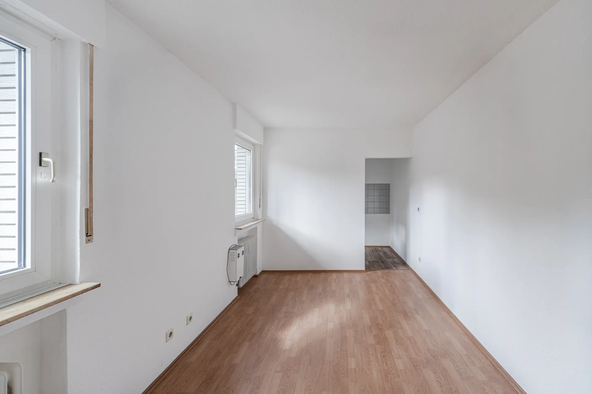 Heller Raum mit weißen Wänden, zwei Fenstern und Laminatboden, Blick in einen angrenzenden Raum.
