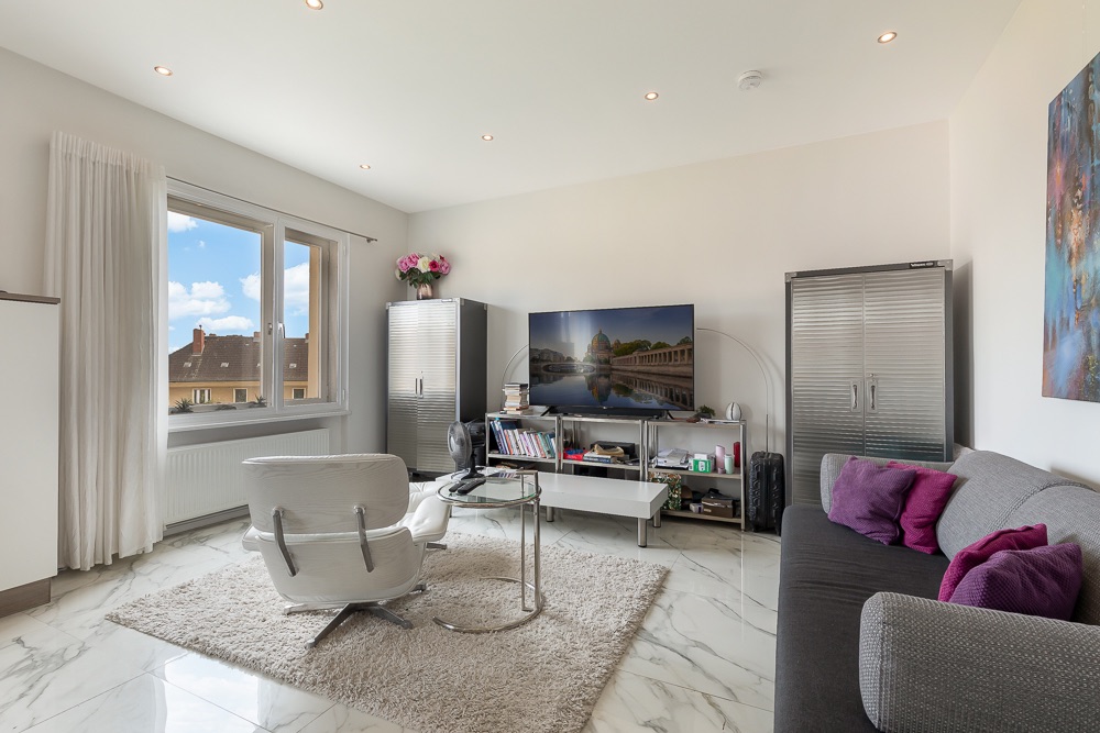 Modernes Wohnzimmer mit grauem Sofa, Sessel, Fernseher und großem Fenster auf weißem Marmorboden.