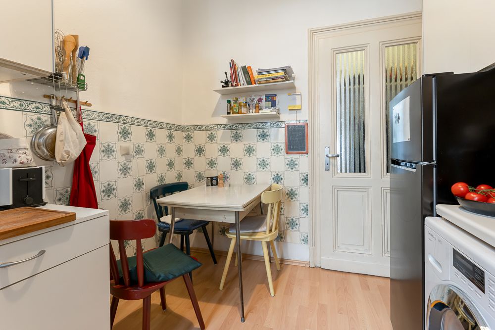 Kleine Küche mit Esstisch, drei Stühlen, Kühlschrank und Wandregal mit Büchern und Gewürzen.