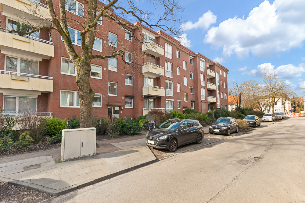 Mehrfamilienhaus aus rotem Backstein mit Balkonen und parkenden Autos an der Straße.