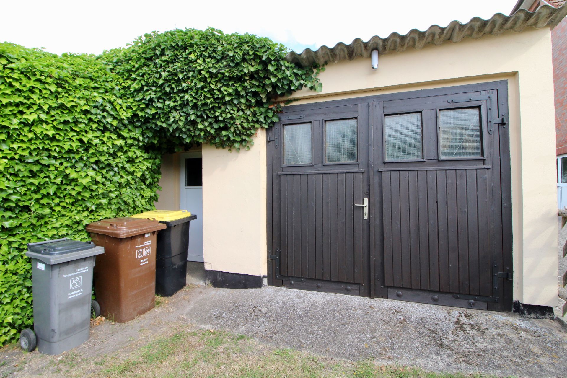 Garage mit braunen Toren, Mülltonnen und grüner Hecke an der Seite.