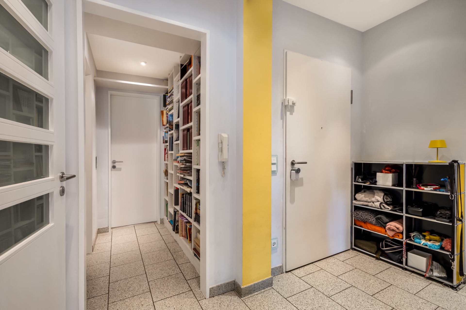 Heller Flur mit Bücherregal, weißer Tür, Regal und gelber Wand.