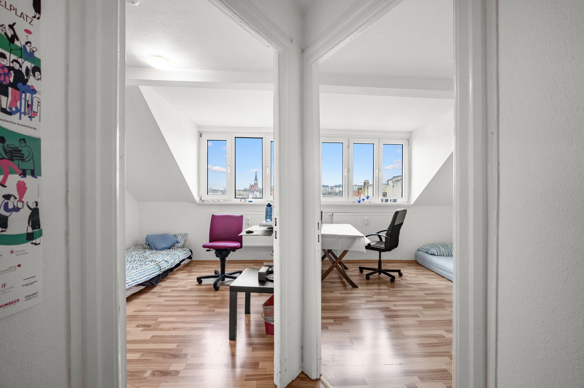 Zwei helle Zimmer mit Schreibtischen, Stühlen, Betten und großen Fenstern mit Blick nach draußen.