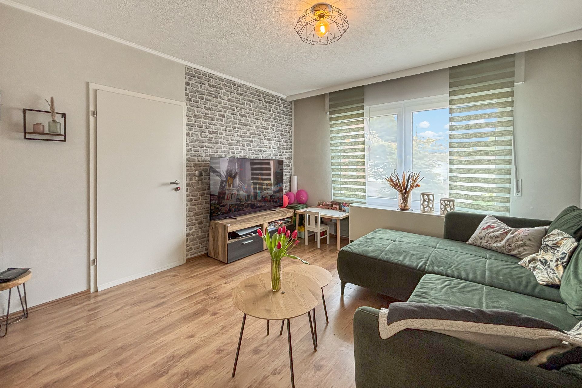 Wohnzimmer mit grünem Sofa, Couchtisch, Fernseher und großen Fenstern mit grünen Jalousien.