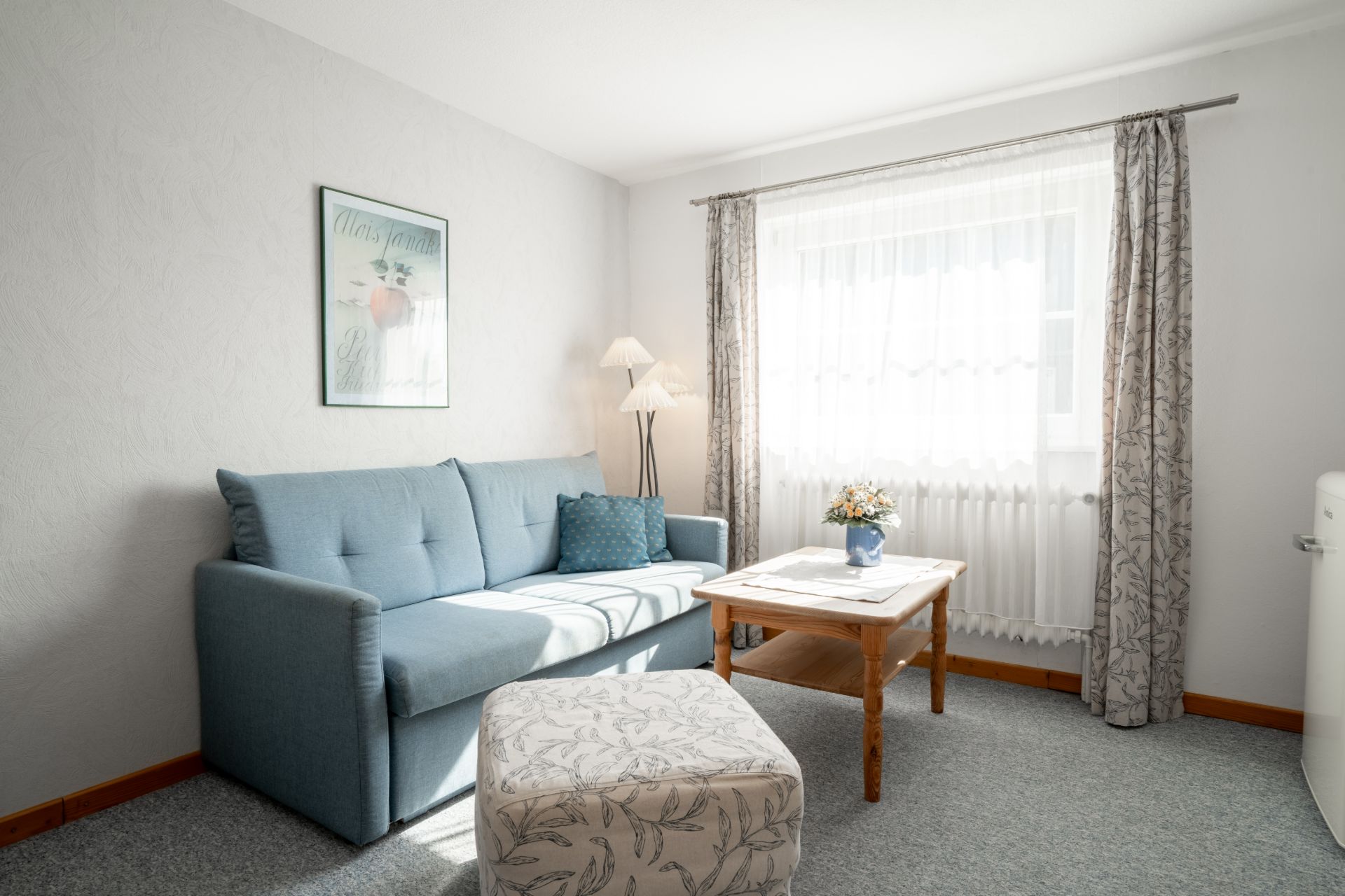 Helles Wohnzimmer mit blauem Sofa, Couchtisch, Hocker und großem Fenster mit Vorhängen.