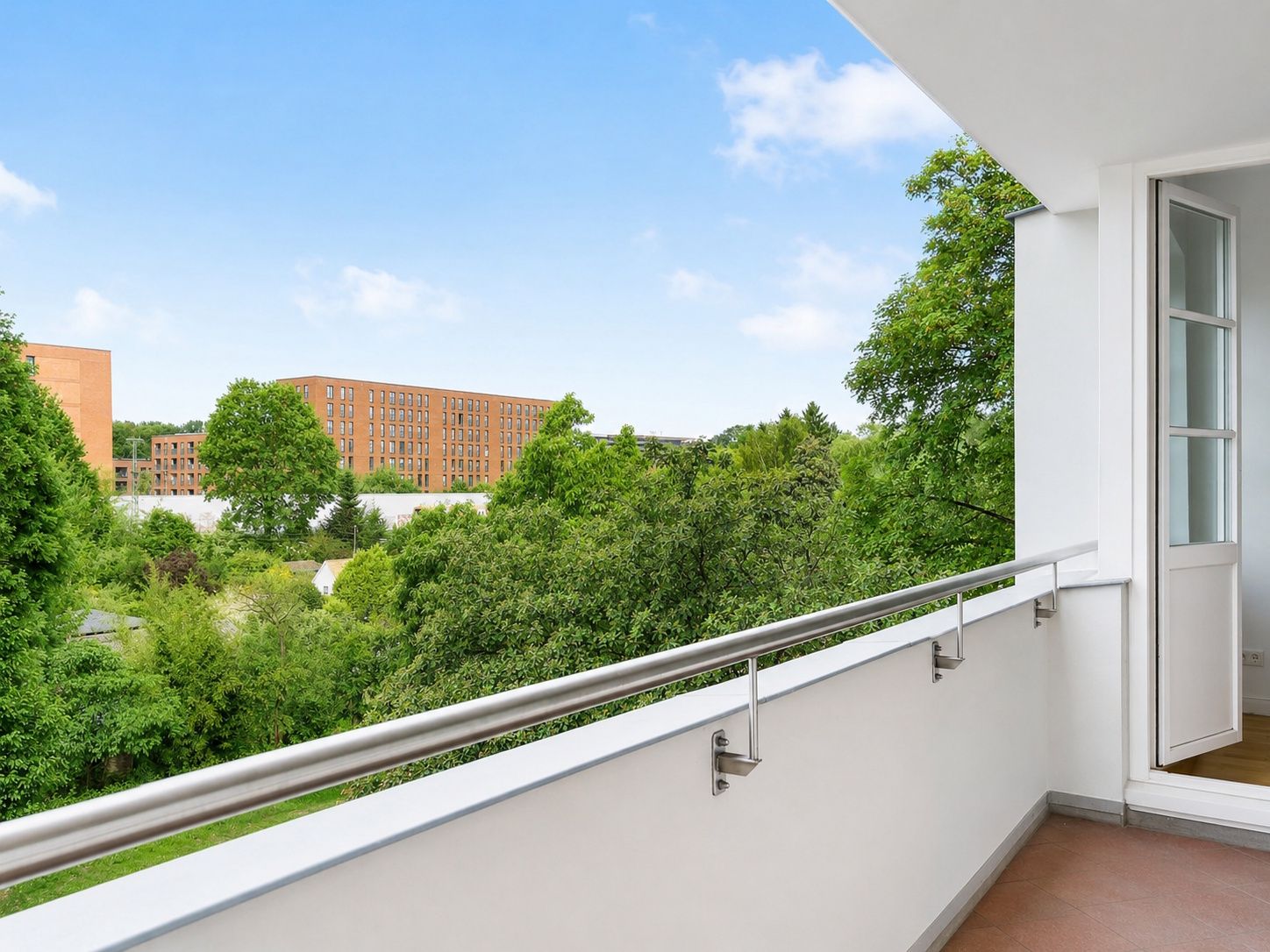 Balkon mit Metallgeländer und Blick auf Bäume und ein rotes Gebäude im Hintergrund.