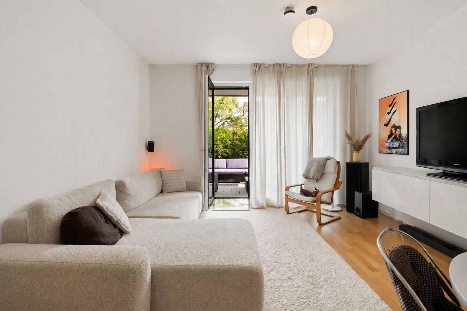 Modernes Wohnzimmer mit heller Couch, Sessel, Fernseher und Zugang zu einem Balkon mit Gartenblick.