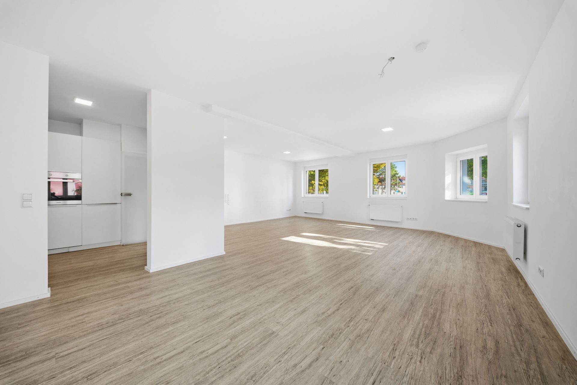Heller, leerer Wohnraum mit Holzfußboden, mehreren Fenstern und einer offenen, weißen Küche.