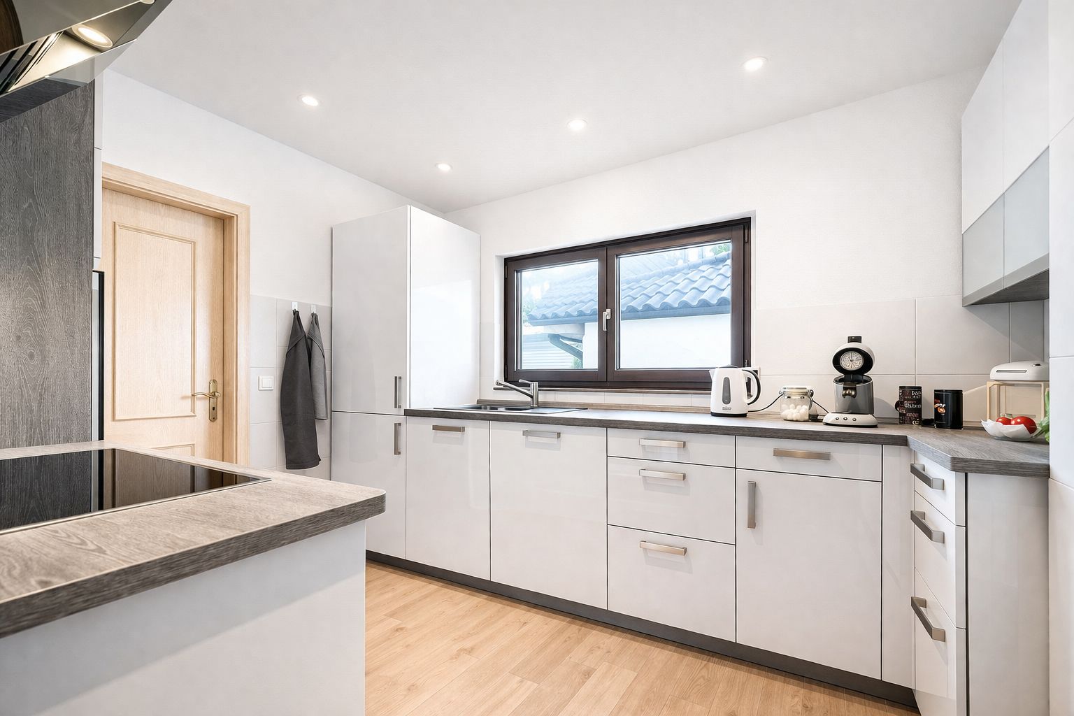 Moderne weiße Küche mit Holzboden, Fenster und verschiedenen Küchengeräten auf der Arbeitsplatte.