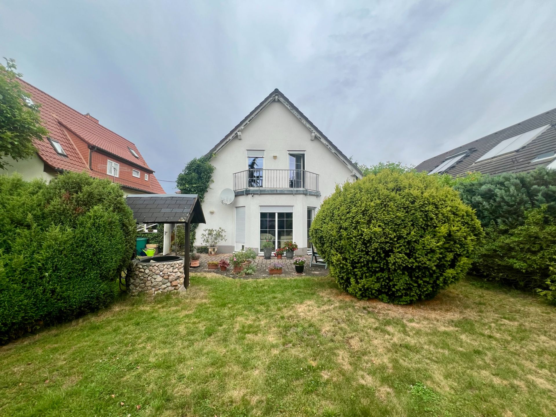 Zweistöckiges Haus mit Balkon und Garten, großer Strauch und Ziehbrunnen im Vordergrund.