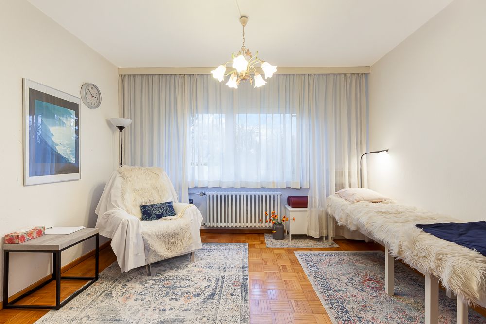 Helles Zimmer mit Sessel, Liege, Teppichen und großem Fenster mit Vorhängen.