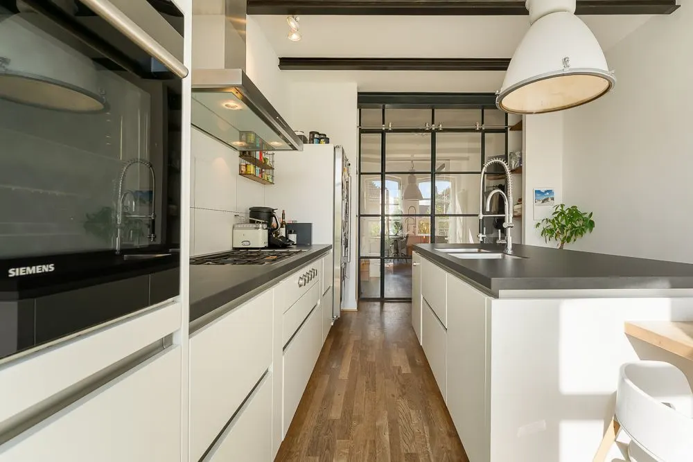 Moderne Küche mit weißer Einrichtung, Holzboden und großem Fenster im Hintergrund.