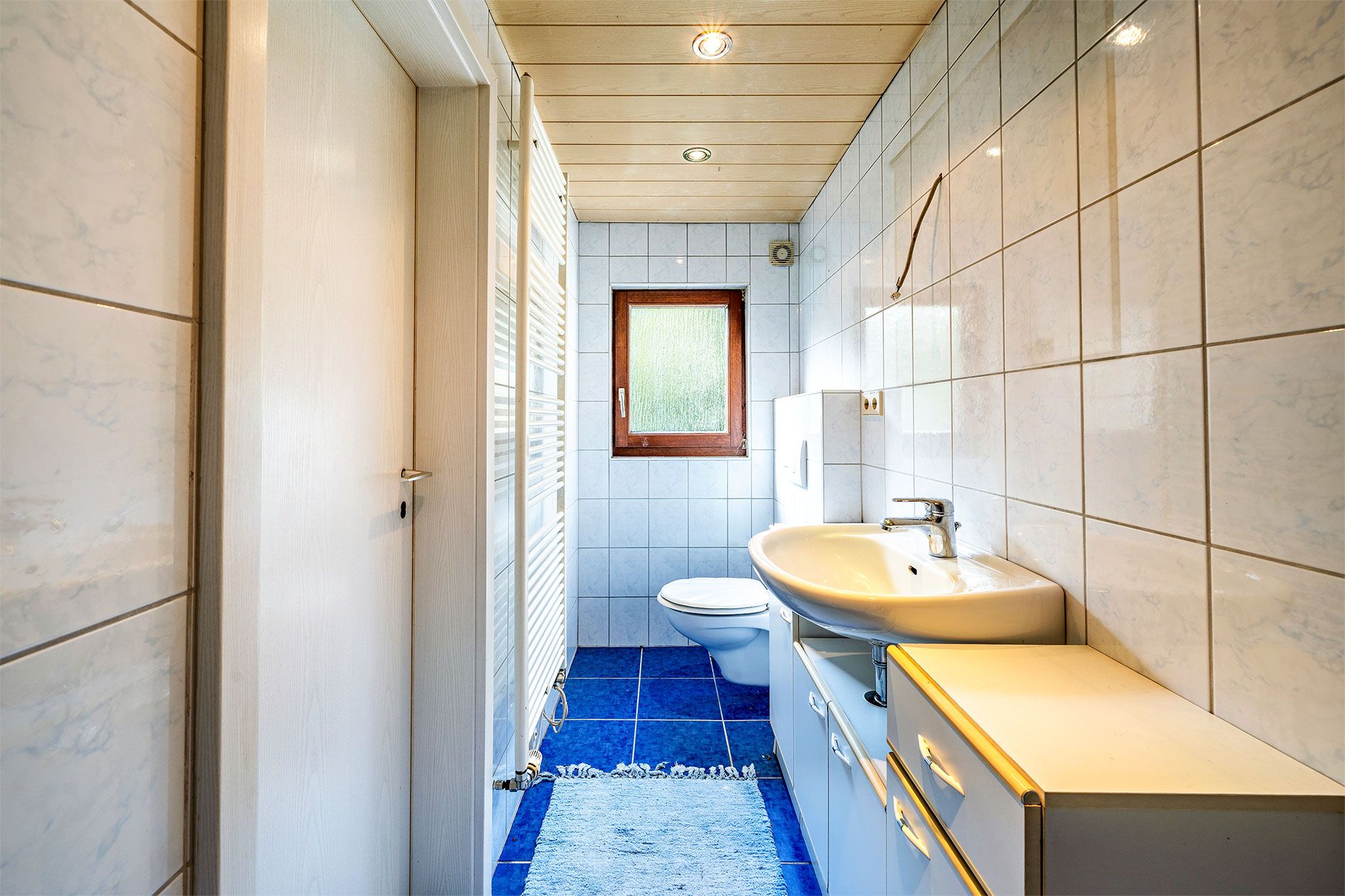 Helles Badezimmer mit blauem Boden, Fenster, Waschbecken, WC und weißen Fliesen.