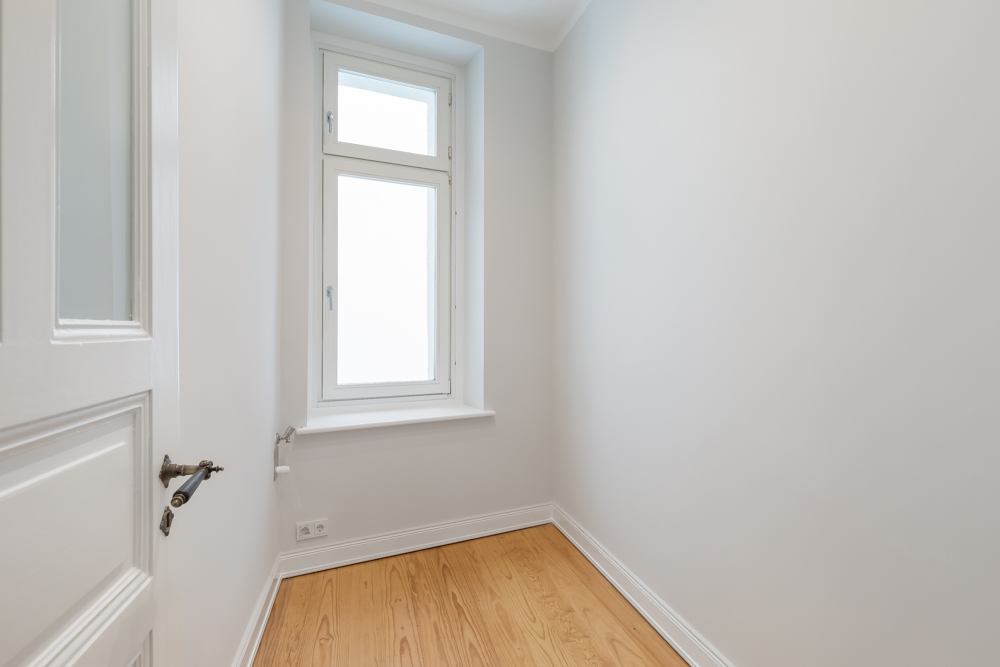 Kleines, leeres Zimmer mit Holzboden und weiß gestrichenen Wänden, Fenster an der Stirnseite.