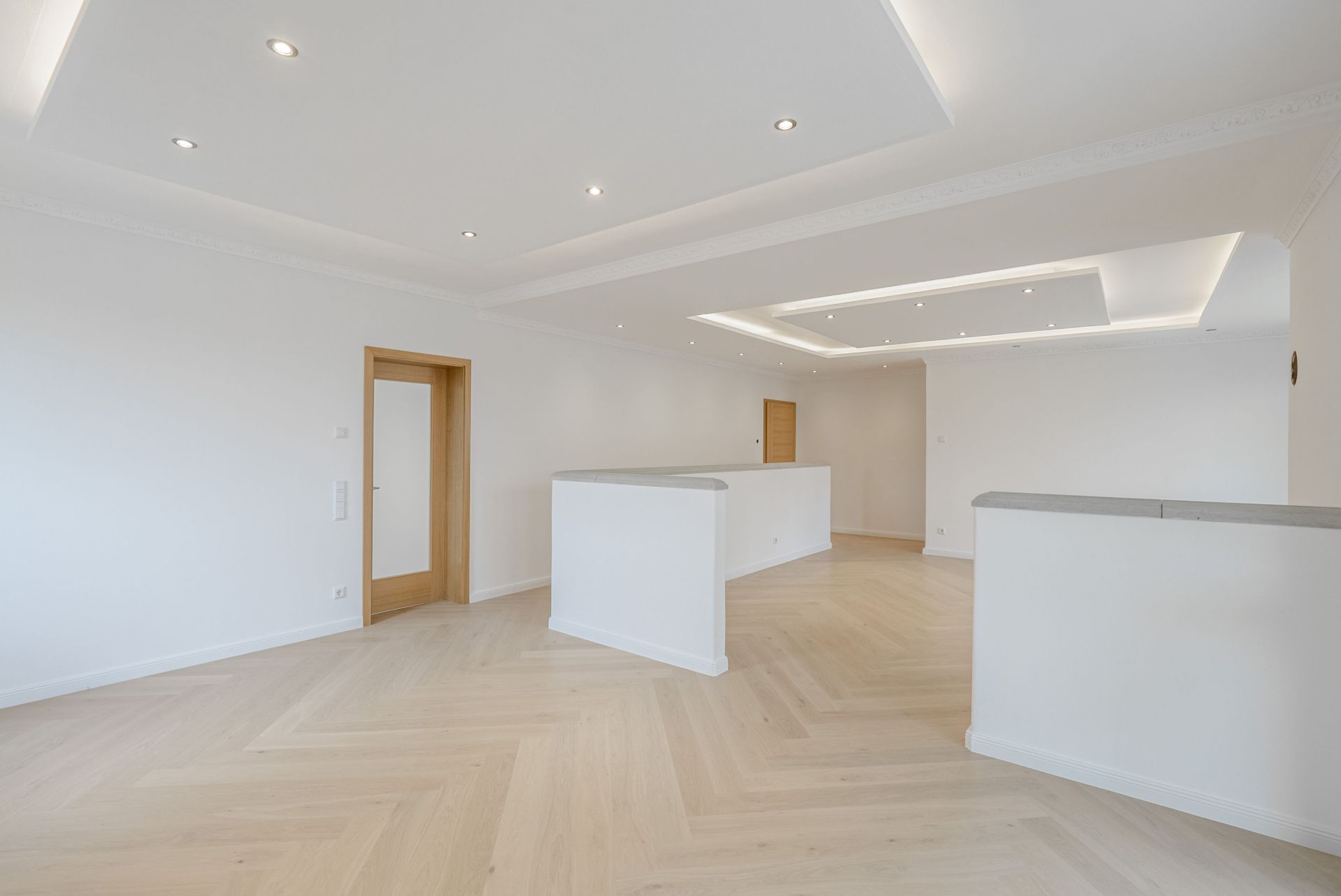 Helles, leeres Zimmer mit Holzfußboden, weißen Wänden und Einbaudeckenleuchten.