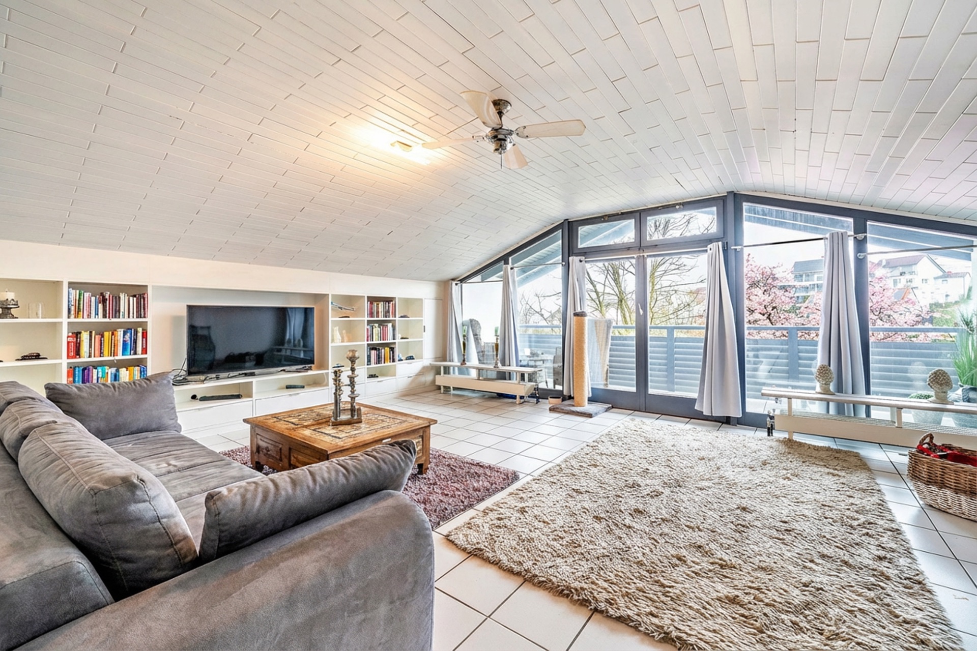 Helles Wohnzimmer mit Sofa, großem Teppich, Couchtisch, Regalwand und großen Fenstern zur Terrasse.