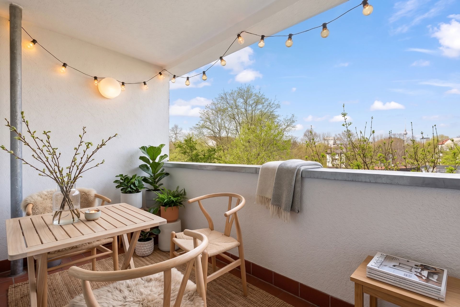 Balkon mit Holztisch, zwei Stühlen, Pflanzen, Lichterkette und Ausblick ins Grüne.