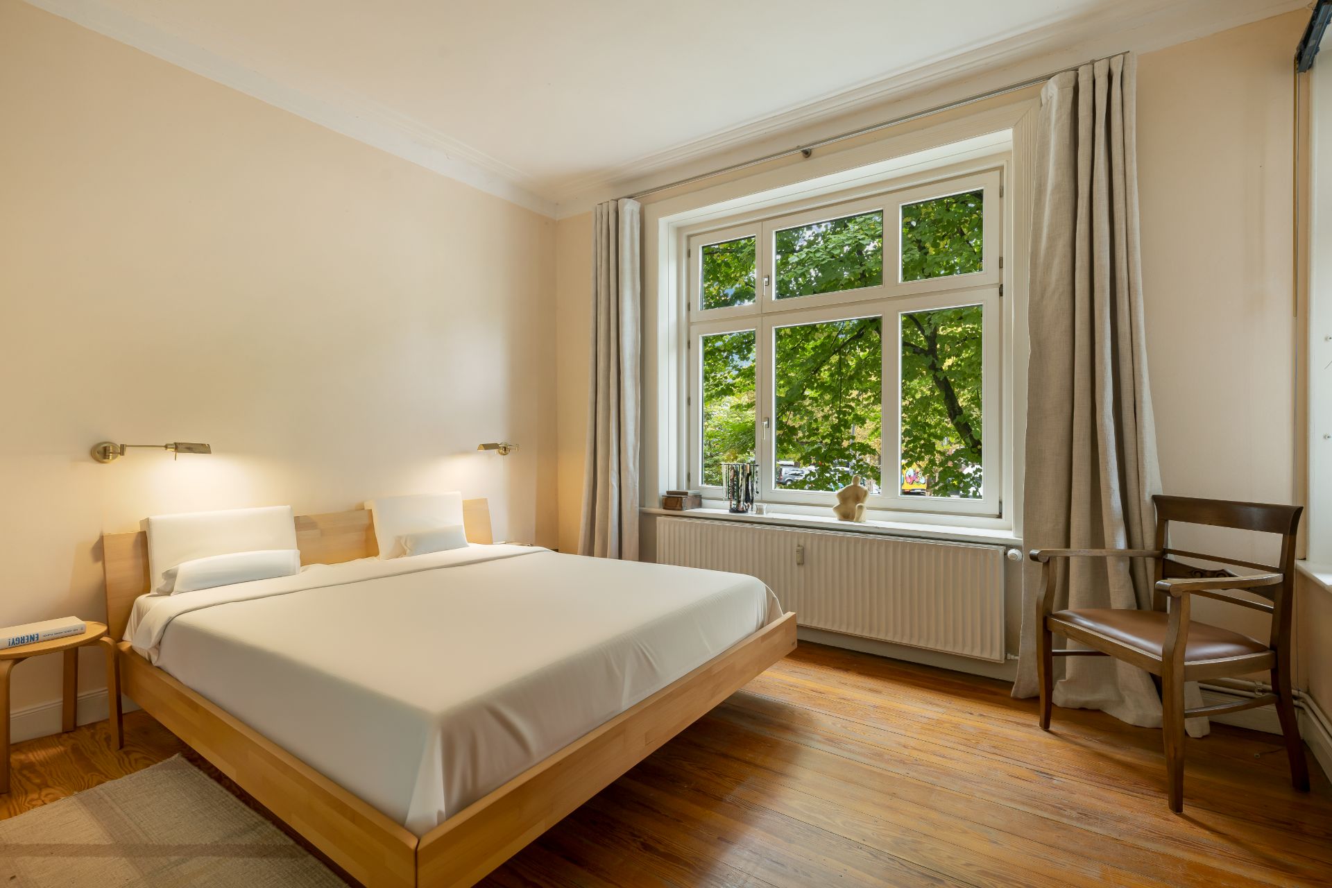 Helles Schlafzimmer mit Doppelbett, Holzfußboden, großem Fenster und zwei Stühlen.